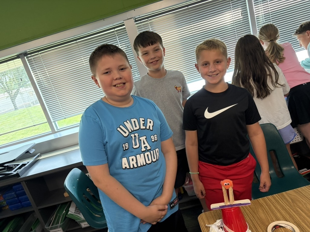 Three boys standing together smiling next to a red cup tower topped with craft sticks and a pom-pom figure.