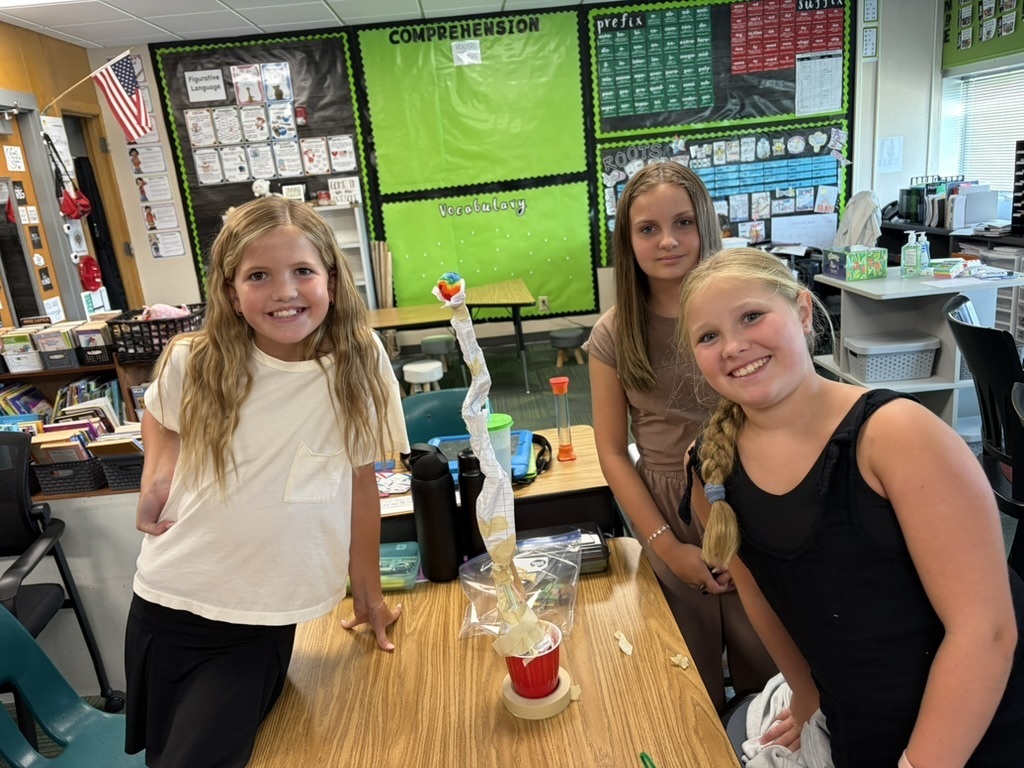 Three girls standing behind a desk presenting a tall vertical structure made of paper and tape attached to a red cup.