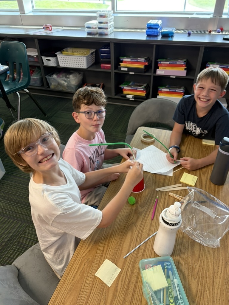 Three boys wearing glasses smiling at a desk while constructing a structure with pipe cleaners, paper, and a red cup.