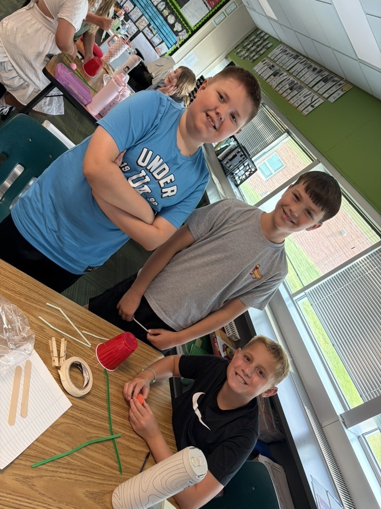 Three boys posing at a classroom desk with craft supplies like straws, tape, and a red cup.