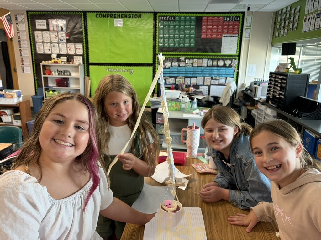 Four girls gathered at a desk showing off a tall paper-and-tape tower project.