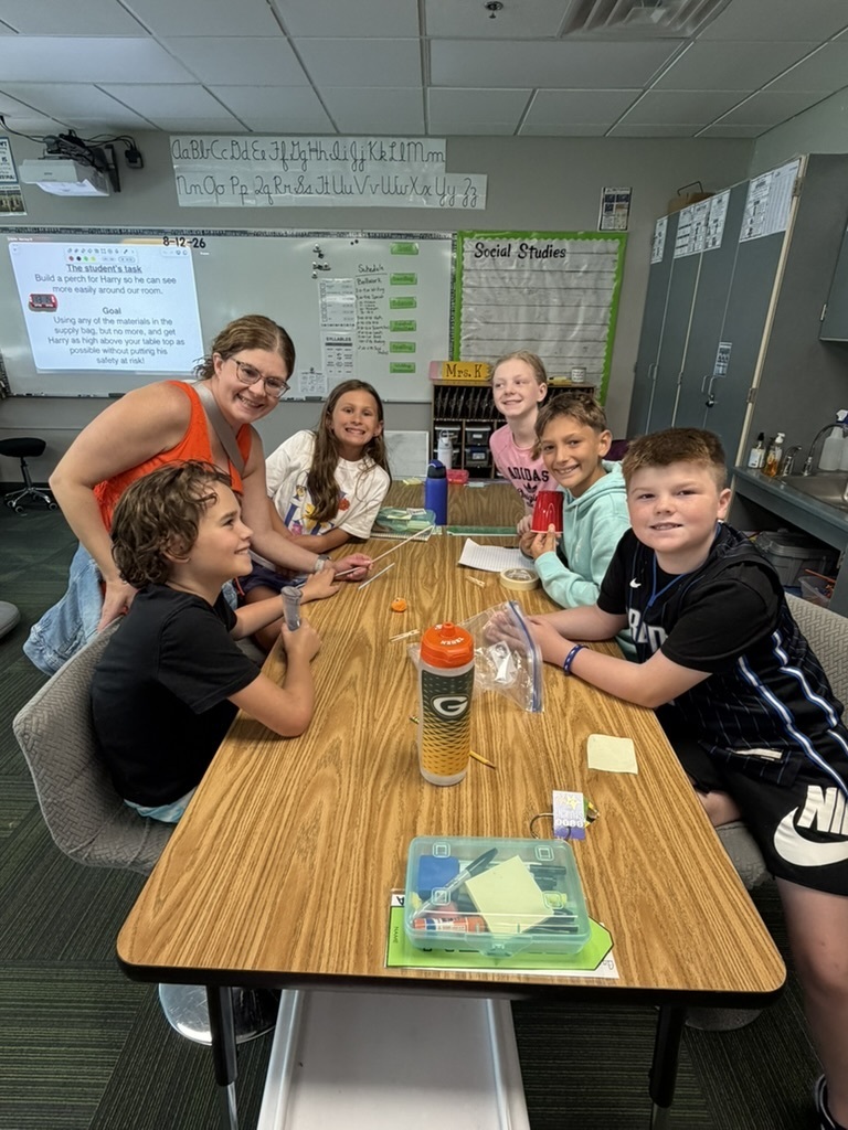 A teacher and five students sitting around a classroom table working on a STEM building activity.
