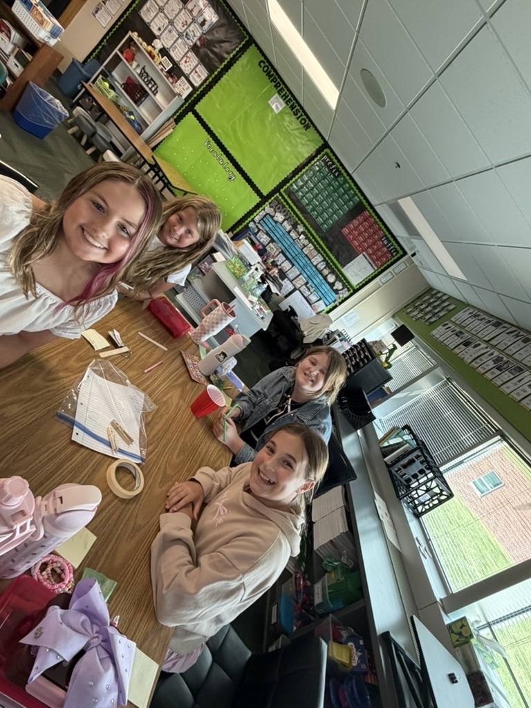 Four smiling girls seated at a long wooden desk in a classroom with project supplies.