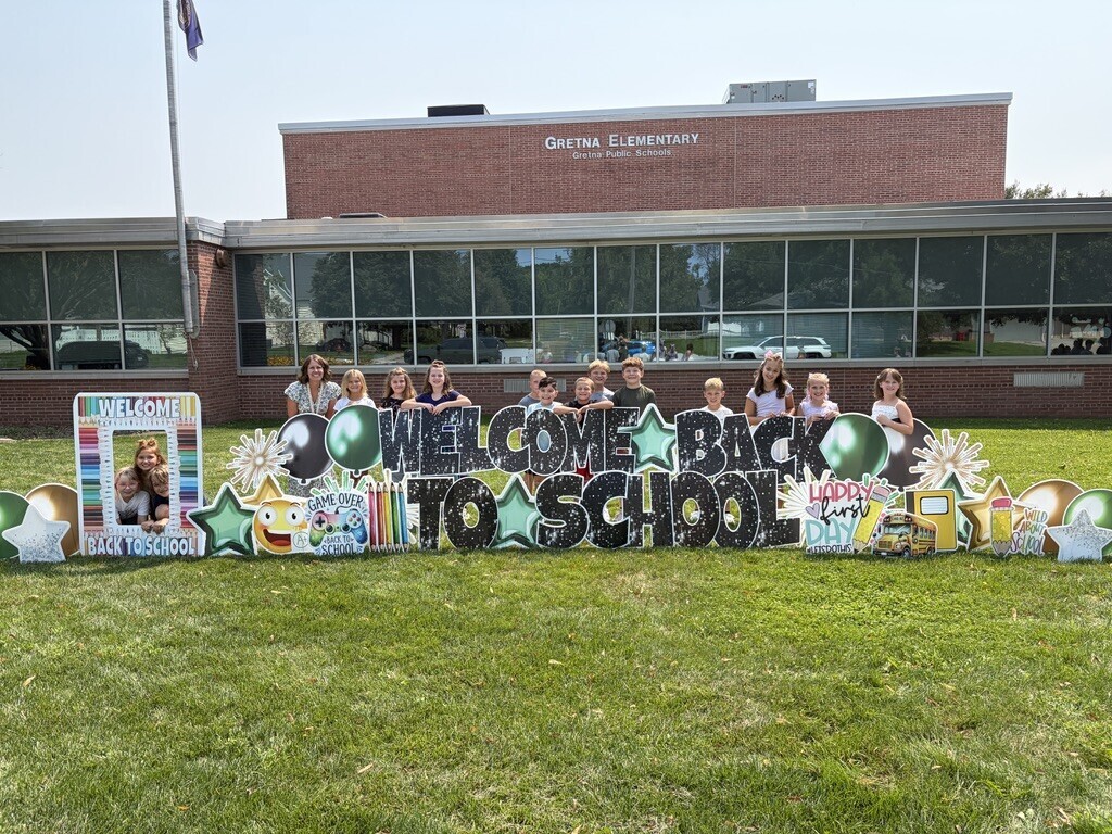 A teacher and a group of young students smiling behind a large "Welcome Back to School" yard sign in front of Gretna Elementary School.