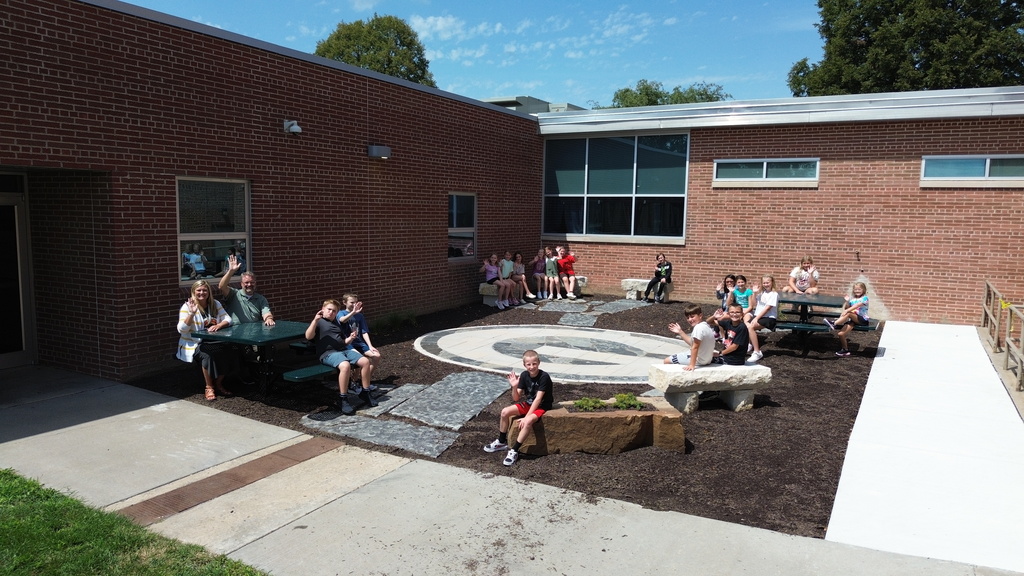 Students and teachers sitting on picnic tables and stone benches, waving in an outdoor courtyard outside a brick school building.