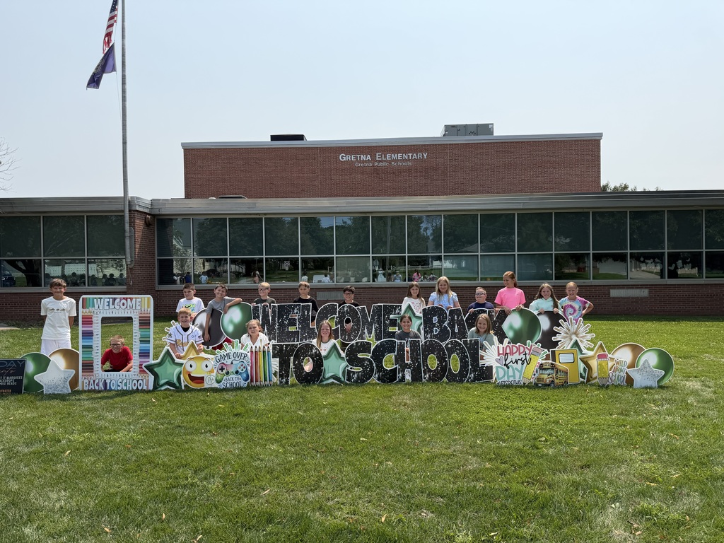 A teacher and a line of young students standing behind a "Welcome Back to School" sign on the front lawn of the school.