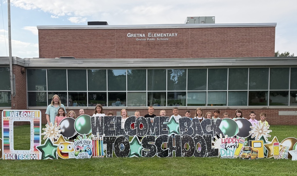 A teacher and a group of young students posing for a photo behind a large "Welcome Back to School" lawn display outside Gretna Elementary School.