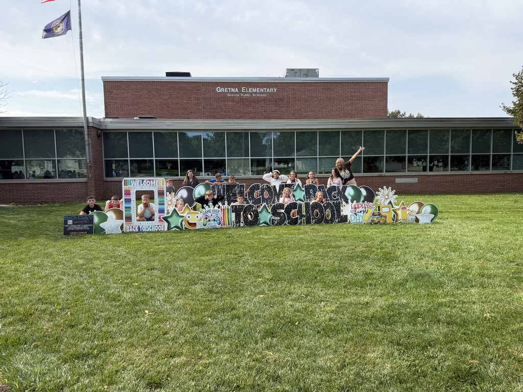 A class of students and their teacher smiling together behind a large "Welcome Back to School" lawn sign outside the school building.