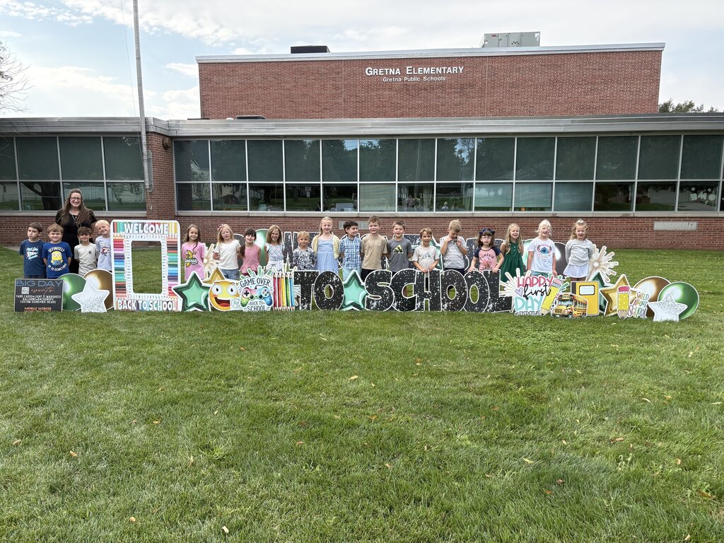 Students and a teacher posing enthusiastically behind a colorful "Welcome Back to School" display outside Gretna Elementary.