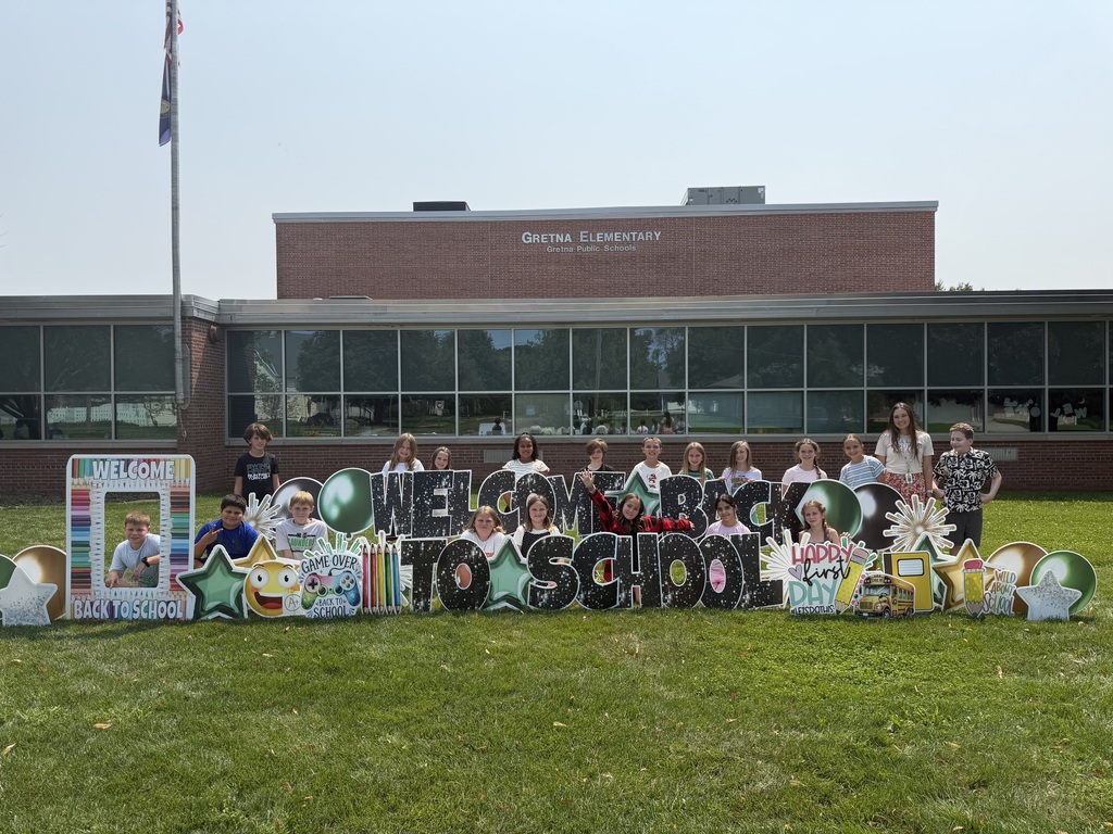 A teacher and a group of elementary school kids standing behind a "Welcome Back to School" sign on the grass in front of Gretna Elementary.