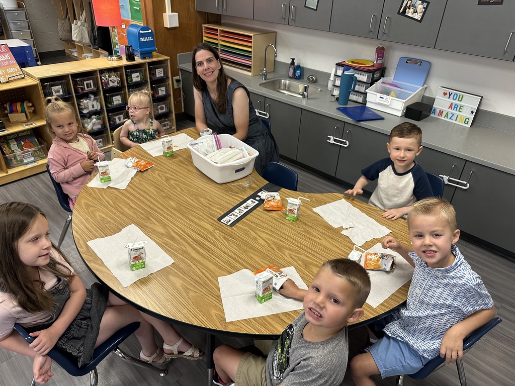 A teacher and six young students sitting around a kidney-shaped wooden table during snack time in a classroom.