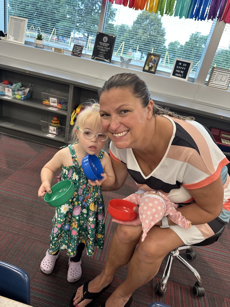 A teacher smiling next to a young girl who is holding colorful plastic play bowls and a baby doll in a classroom.
