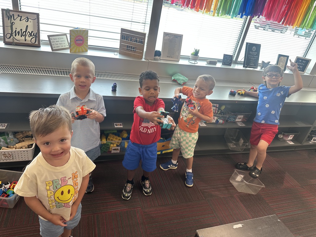 Five young boys smiling and playing together with toy action figures and cars in a classroom setting.