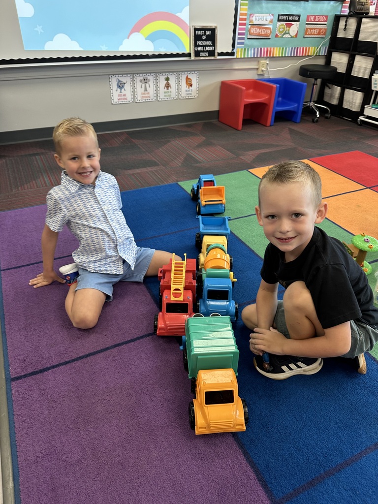 Two young boys sitting on a colorful carpet in a classroom, smiling next to a long line of toy trucks and construction vehicles.
