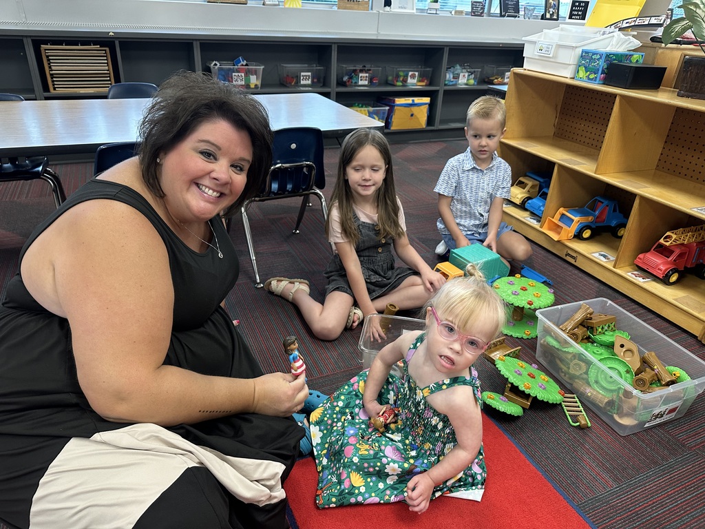 A smiling teacher sitting on the floor in a preschool classroom while playing with toys alongside three young students.