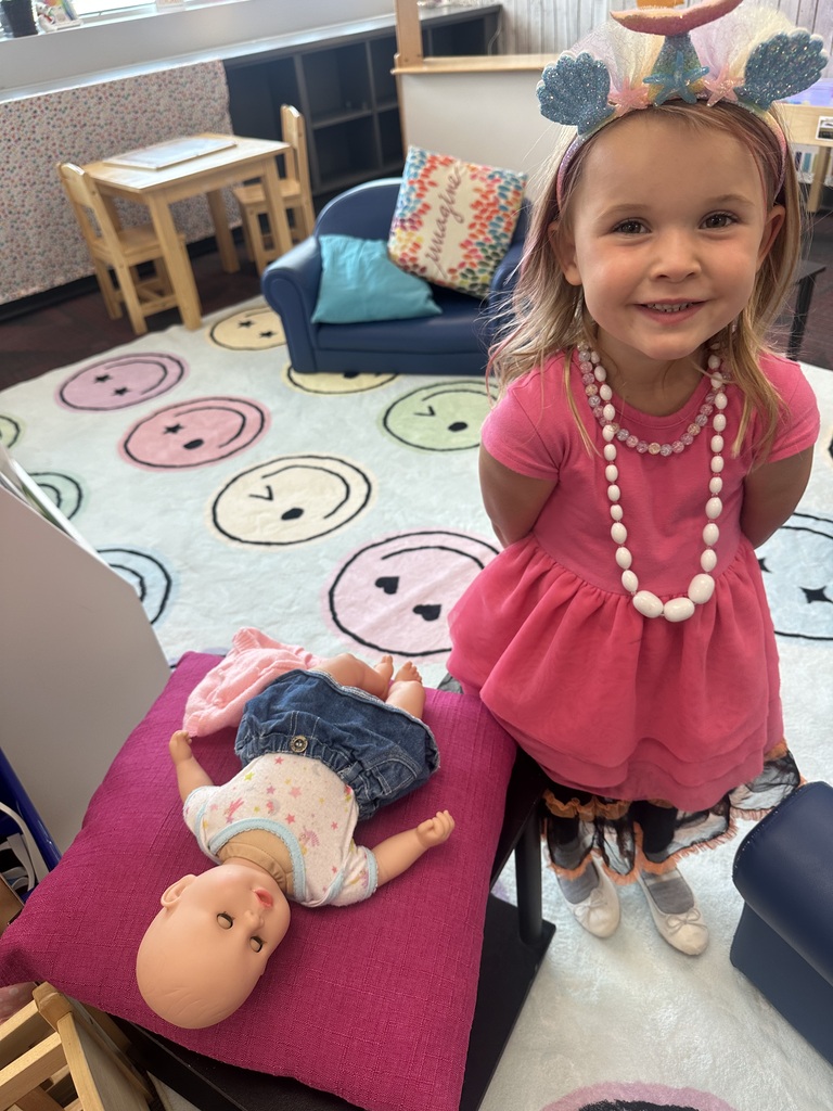 A young girl in a pink dress and mermaid headband smiling in a classroom next to a toy baby doll on a cushion.