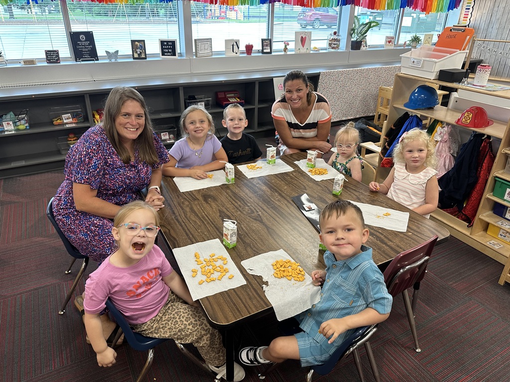 Two adults and six children sitting around a classroom table smiling during snack time with fish crackers and juice boxes.