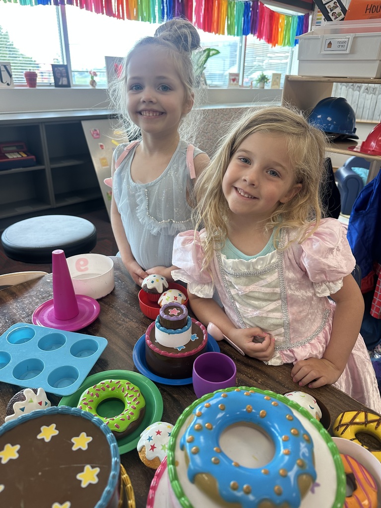 Two smiling young girls posing at a play table filled with colorful toy cakes, donuts, and play food items.