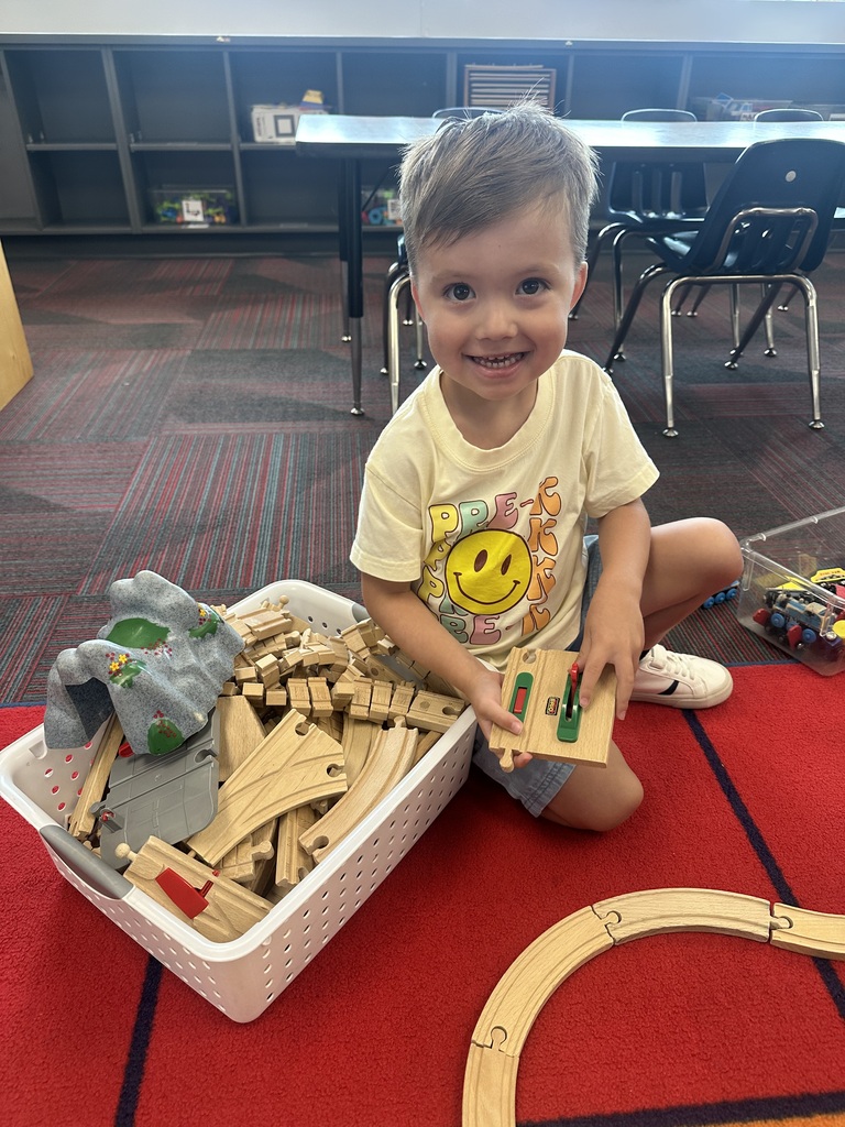 A smiling boy sitting on the floor next to a bin of wooden train track pieces and toy train accessories.
