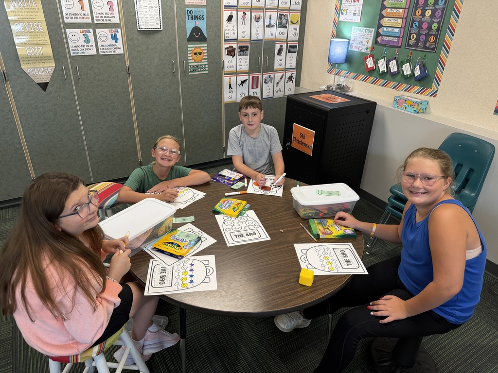 Four students sitting at a round classroom table coloring worksheets with crayons and markers.