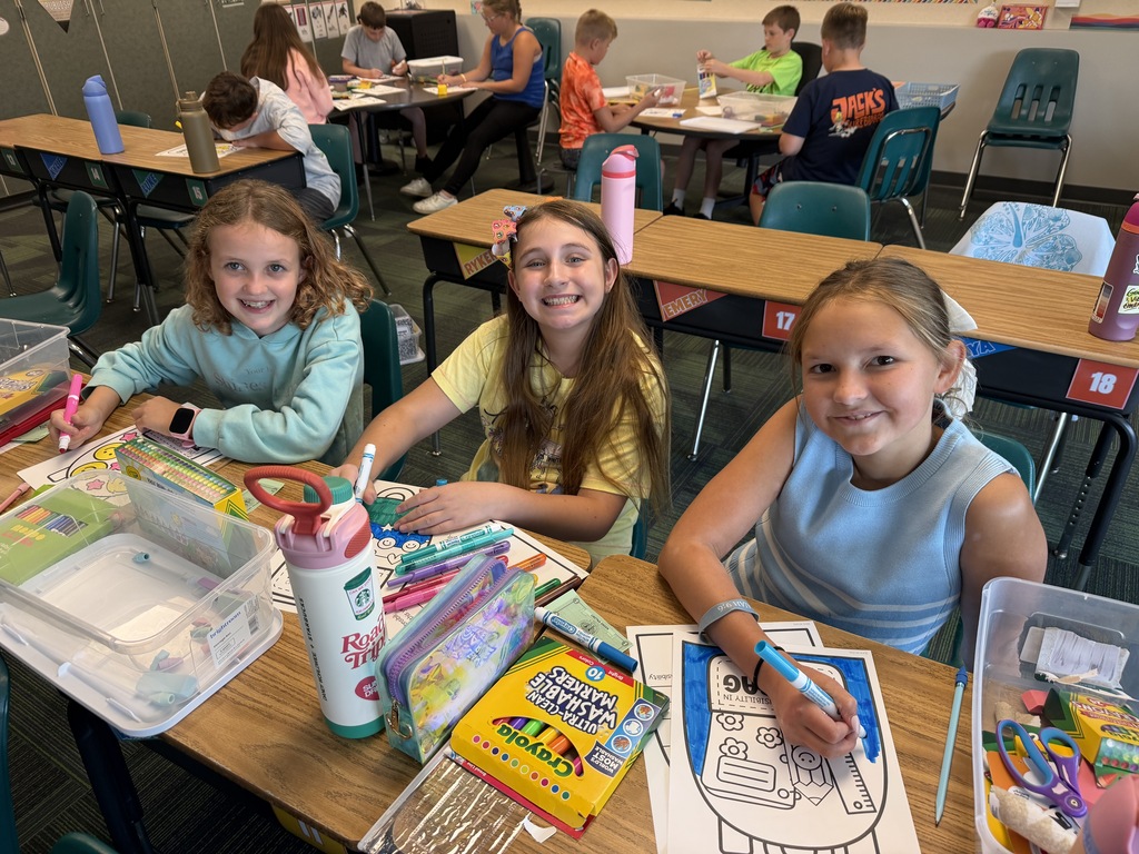 Three elementary school girls smiling at their desks while working on coloring pages with markers and crayons.