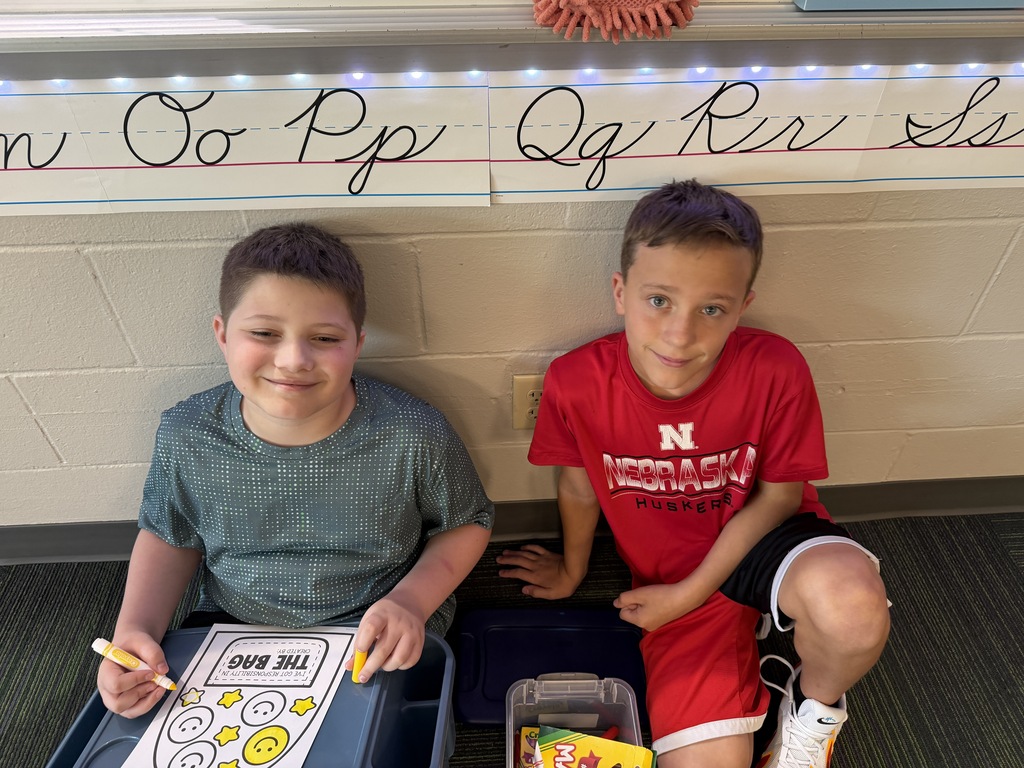 Two boys smiling while sitting on the floor together against a wall with cursive alphabet posters above them.