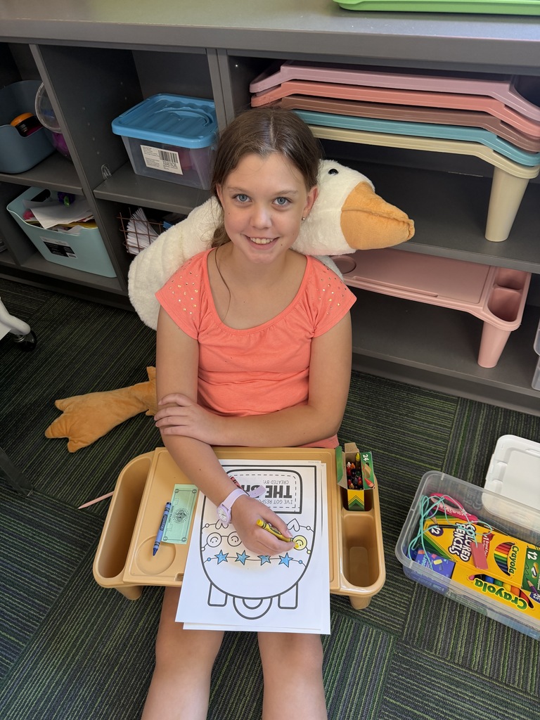 A smiling girl sitting on the floor with a plush duck behind her, coloring a worksheet on a lap desk.