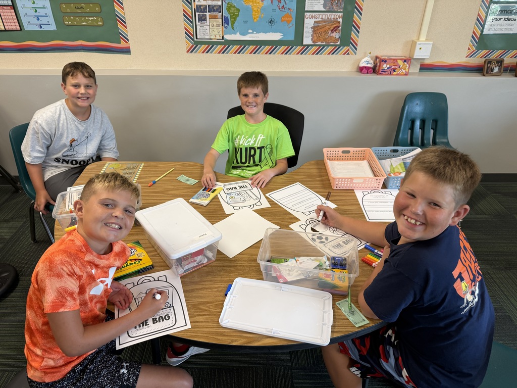 Four students sitting at a round classroom table coloring worksheets with crayons and markers.