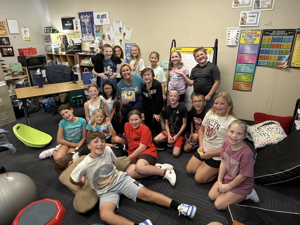 A teacher holding a children's book and posing for a group photo with a room full of smiling elementary students.