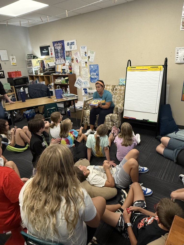 A teacher sitting in an armchair showing the cover of a children’s book to a group of students sitting on the classroom floor.