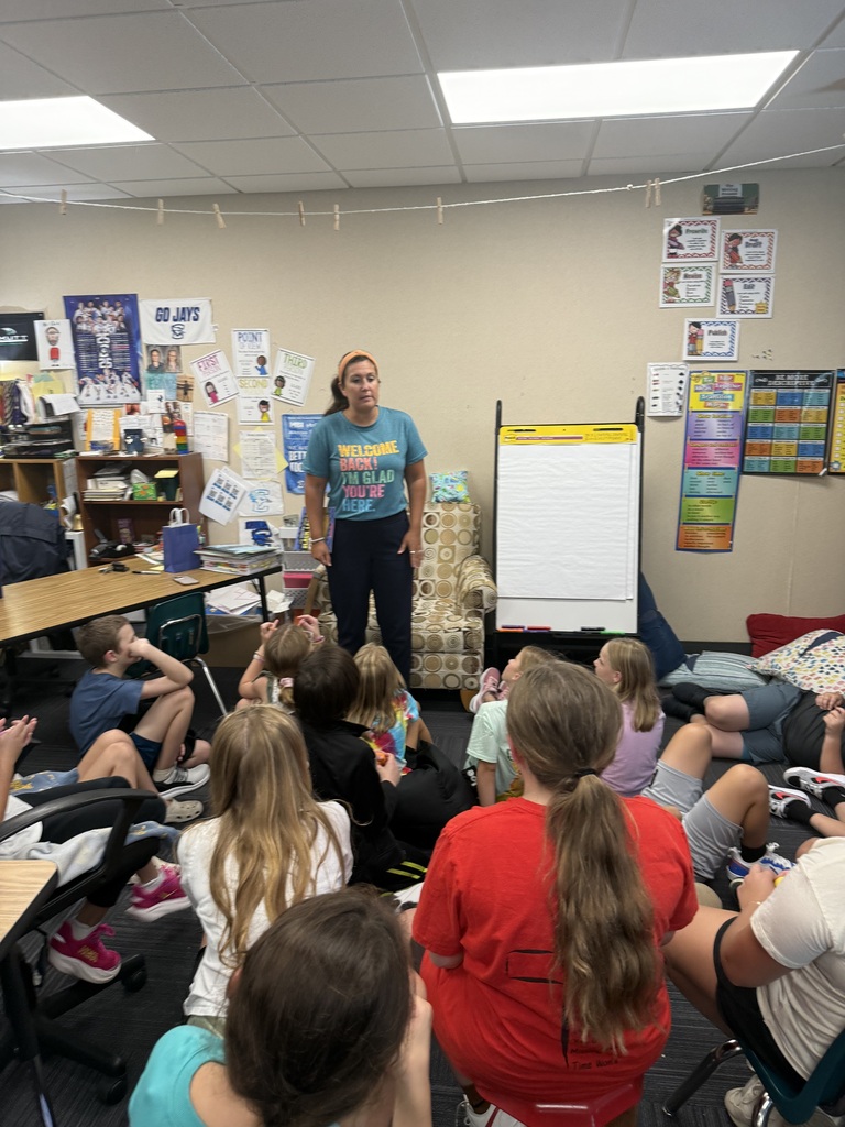 A teacher in a teal shirt standing in front of a classroom of students seated on the floor listening.