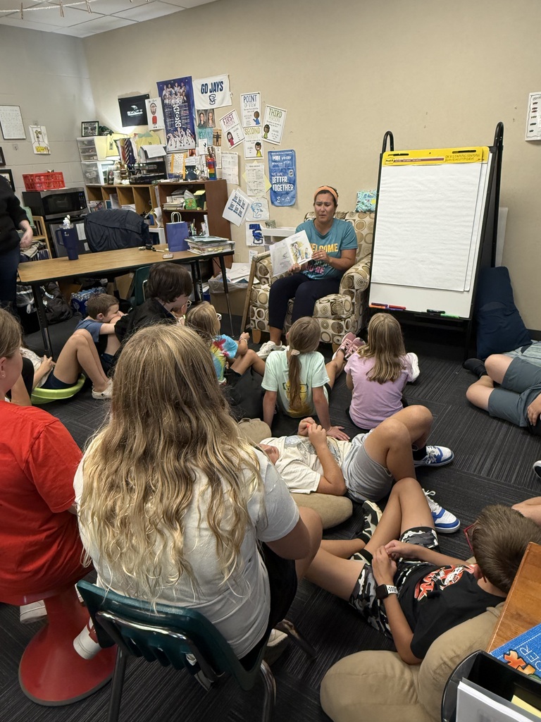 A teacher sitting in an armchair reading an open children's book aloud to students gathered on the carpet.