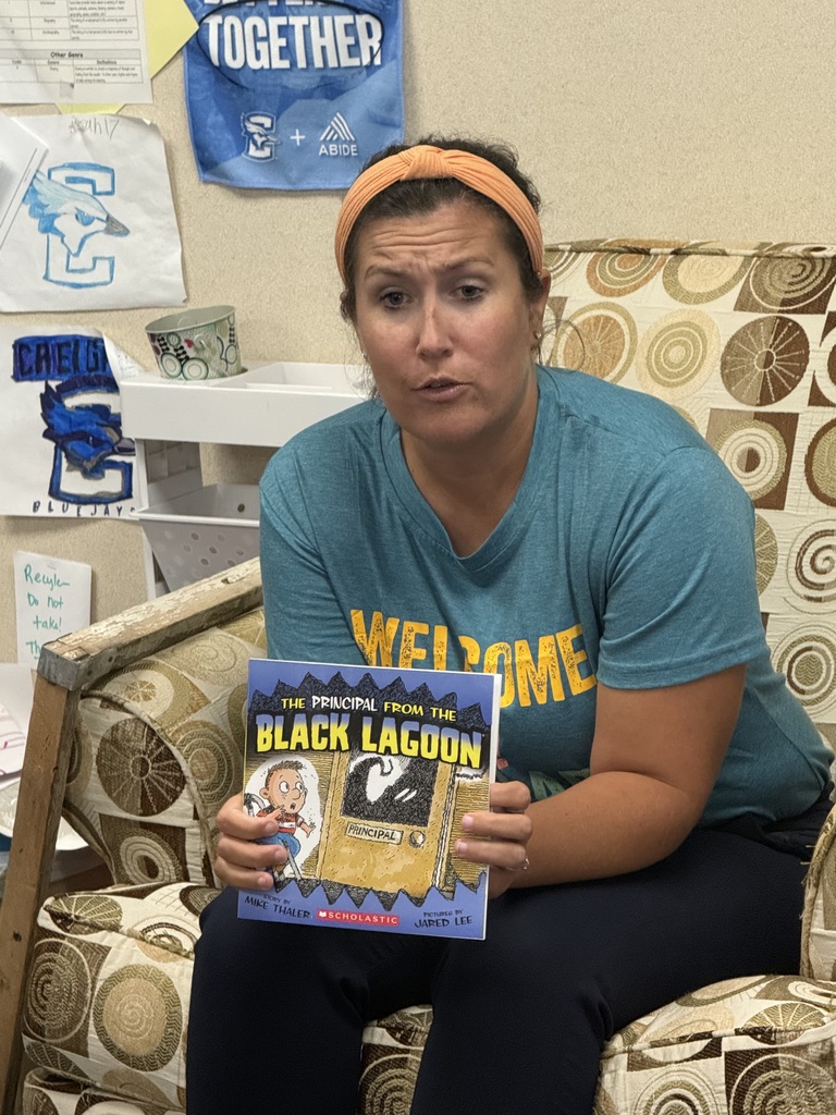 A close-up of a teacher seated in an armchair holding up the book The Principal From the Black Lagoon.