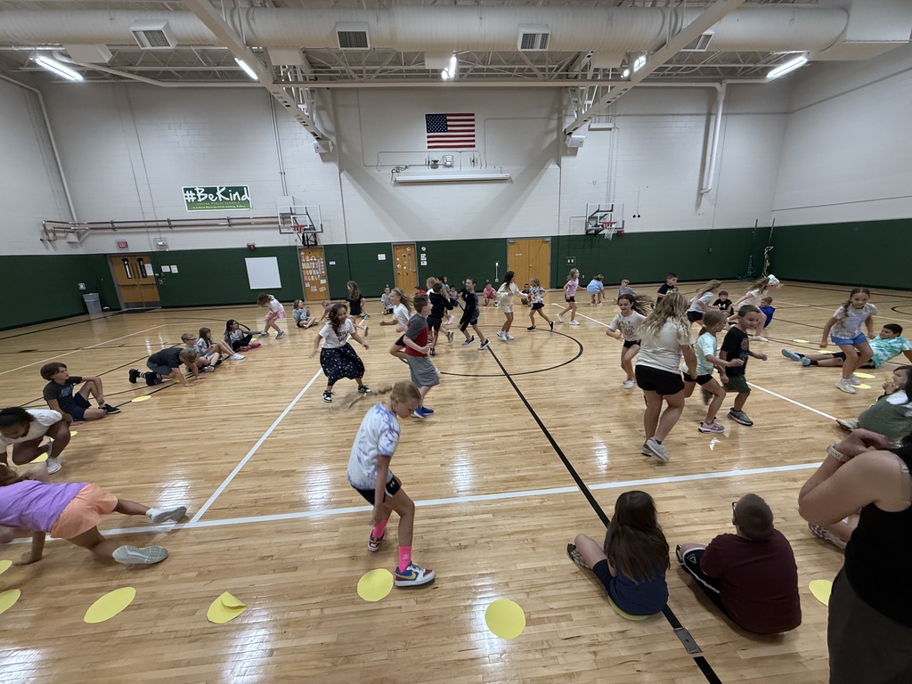 Children scattered across a wooden gymnasium floor, running and playing an interactive activity near yellow markers.