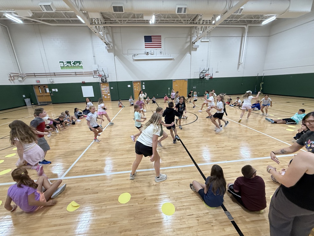 Students actively running around yellow floor markers during a game on a gym court while others watch from the perimeter.