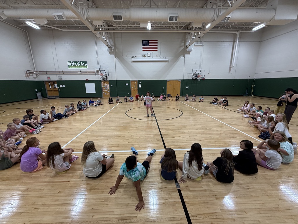 A wide view of a school gymnasium where a large group of students sits in a wide circle around the basketball court as one student stands in the center.
