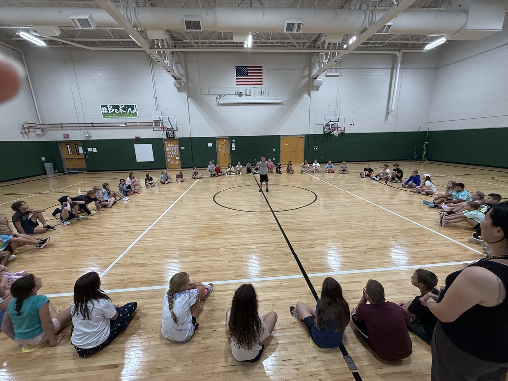A wide shot of students seated in a large circle on the gym floor listening as a young boy stands in the middle circle to speak.