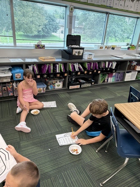 Two students sitting on a classroom carpeted floor, working on worksheets next to small bowls of snacks.