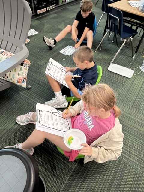 Three students sitting on flexible floor chairs in a classroom, filling out worksheets on clipboards.