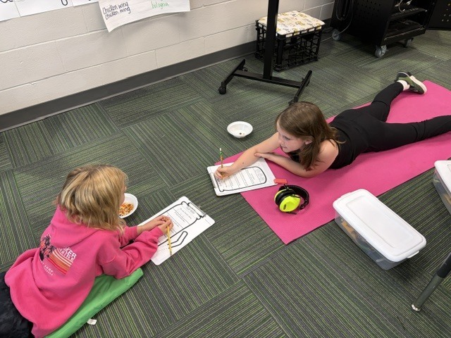 Two young girls lying on the carpeted floor opposite each other, writing on clipboards.