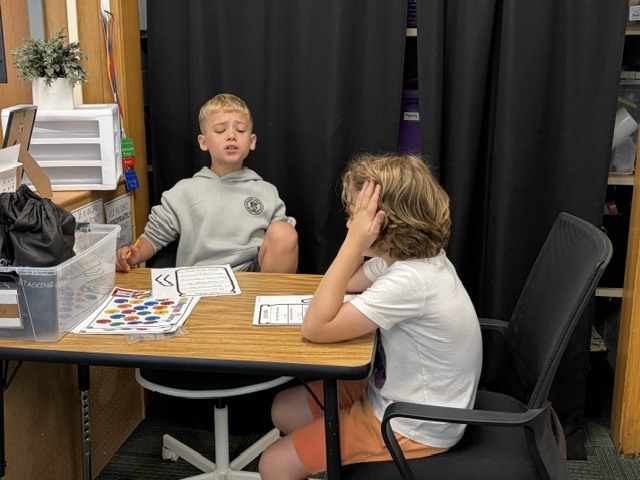 Two students sitting opposite each other at a small classroom desk, working on worksheets together.