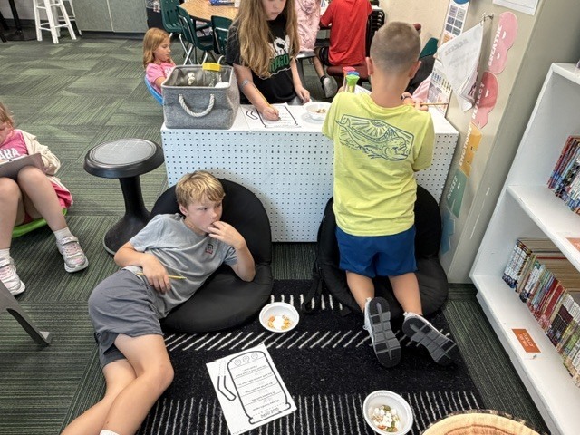Elementary students working in a classroom area with flexible seating, books, and low tables.