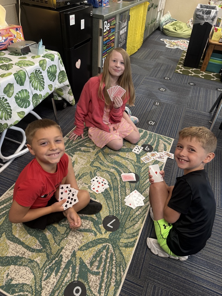 Three students sitting on a green botanical-patterned rug in a classroom, smiling and holding playing cards.