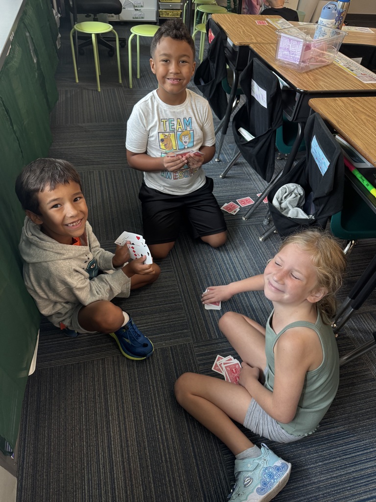 Three students sitting on a dark grey carpet in a classroom, smiling while holding playing cards.
