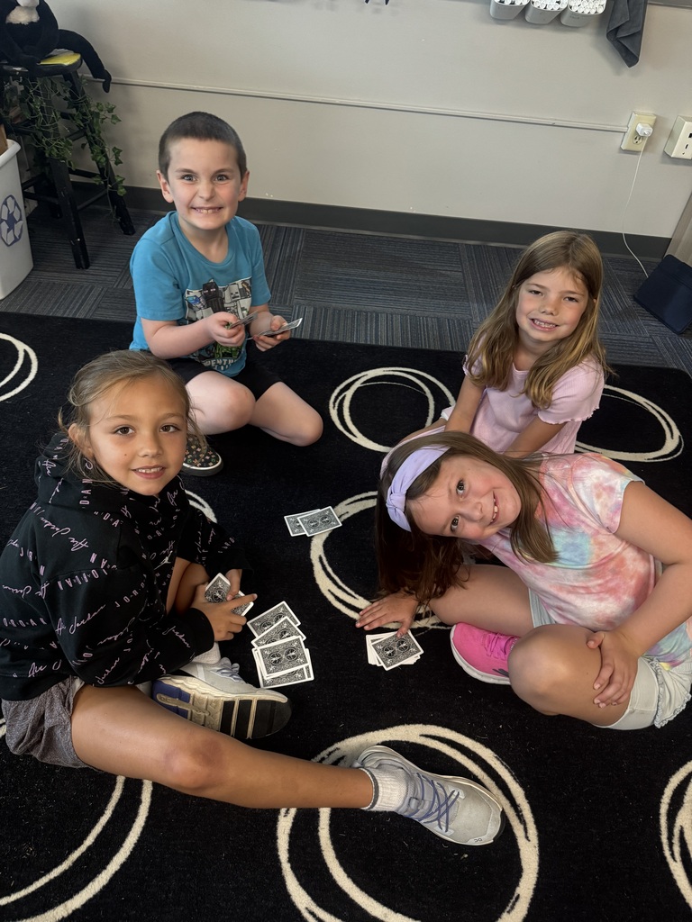 Four young students sitting on a black patterned rug in a classroom, smiling while holding and playing with cards.