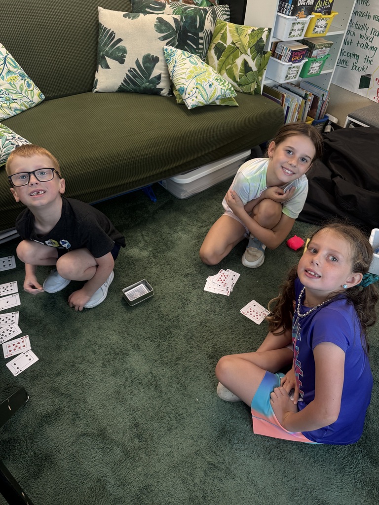 Three young students sitting on a green rug near a couch in a classroom, smiling while playing a card game.