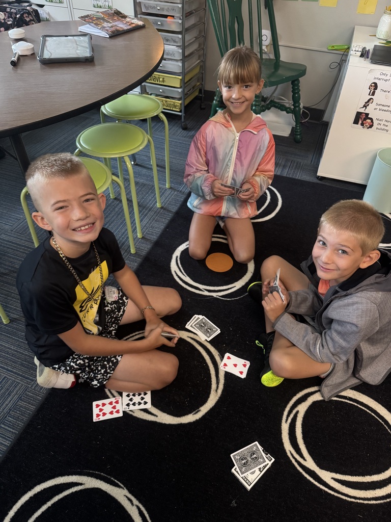 Three young students sitting on a black patterned rug in a classroom, smiling as they play a card game.