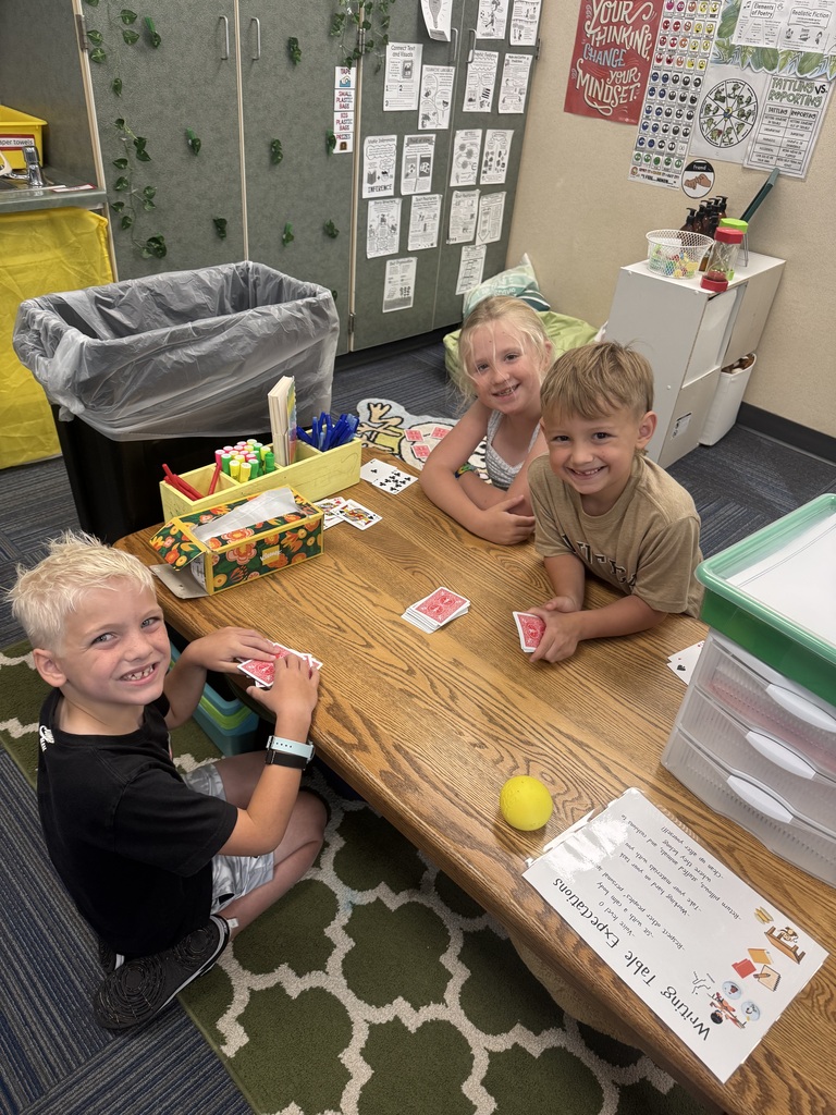 Three young students gathered around a wooden activity table in a classroom, holding cards and smiling.