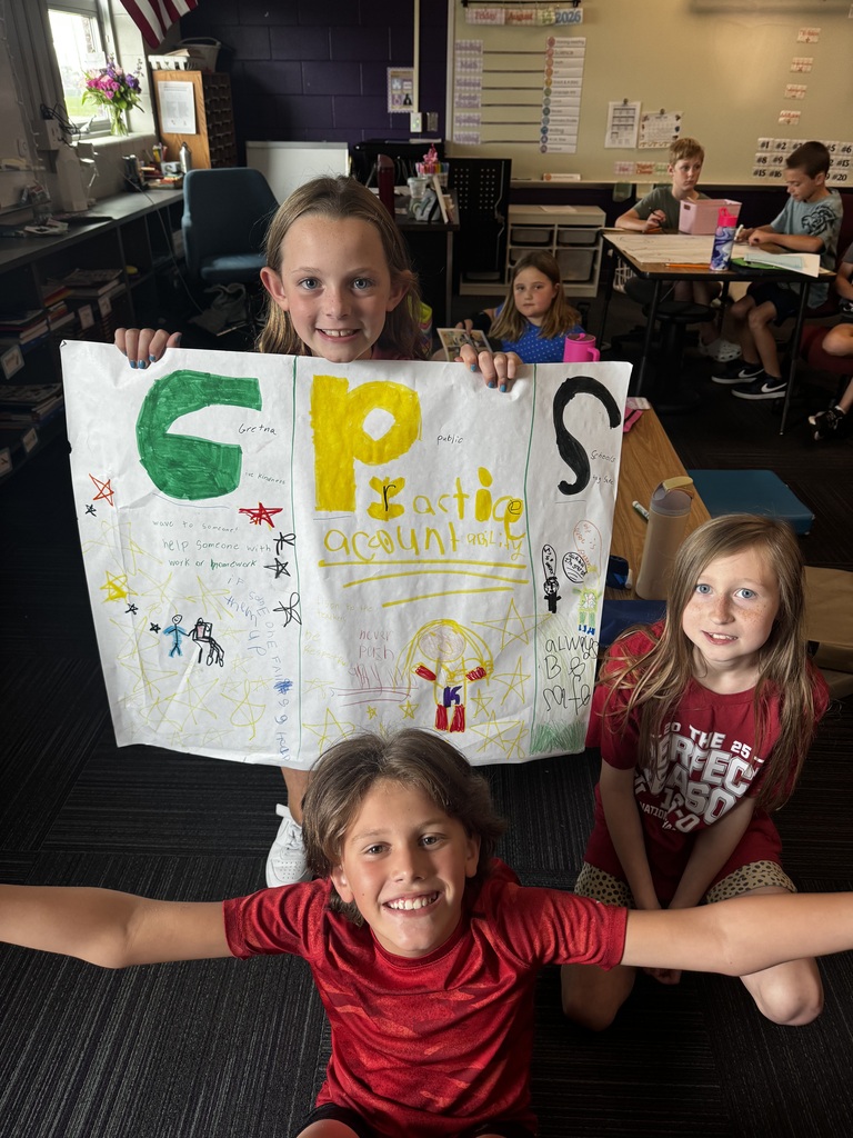 Three children pose with a brightly colored, hand-drawn "GPS" poster in a classroom setting, with one boy kneeling in front with his arms outstretched.