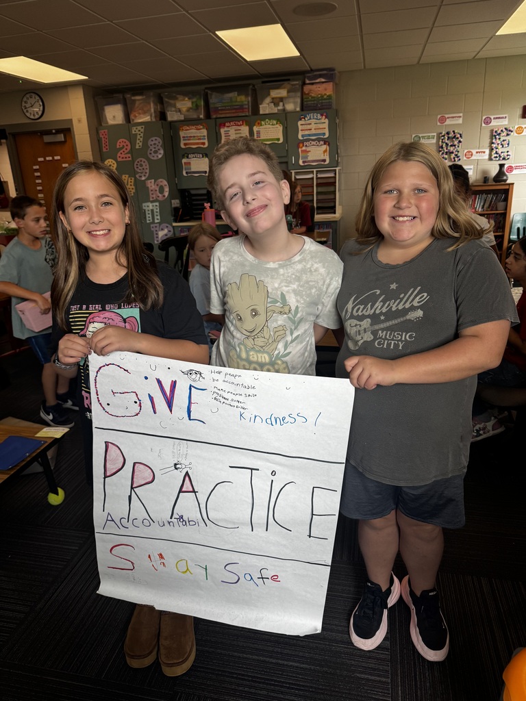 Three smiling students in a classroom display a large white poster with colorful, handwritten text that reads "Give Kindness, Practice Accountability, Stay Safe."